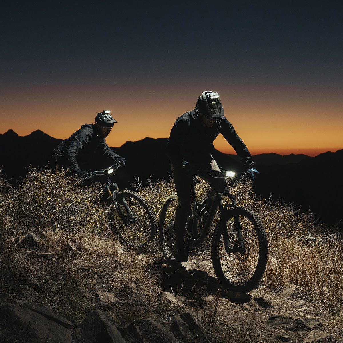 Two mountain bikers descending a ridge at dusk with their bike lights on
