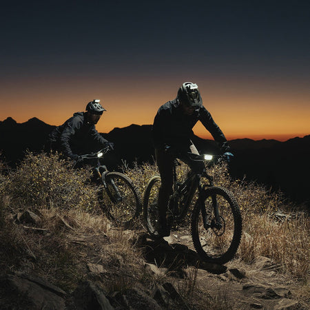Two mountain bikers descending a ridge at dusk with their bike lights on