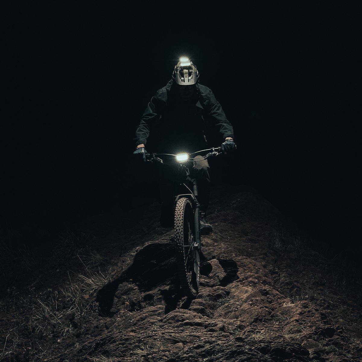 Mountain biker riding toward the camera on a dark trail lit by handlebar and helmet bike lights